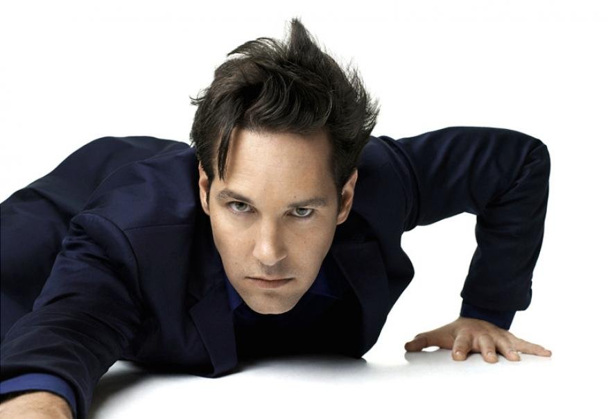 Actor Paul Rudd lies prone on a white surface in a navy suit jacket with an intense gaze directed at the camera in a portrait shot.