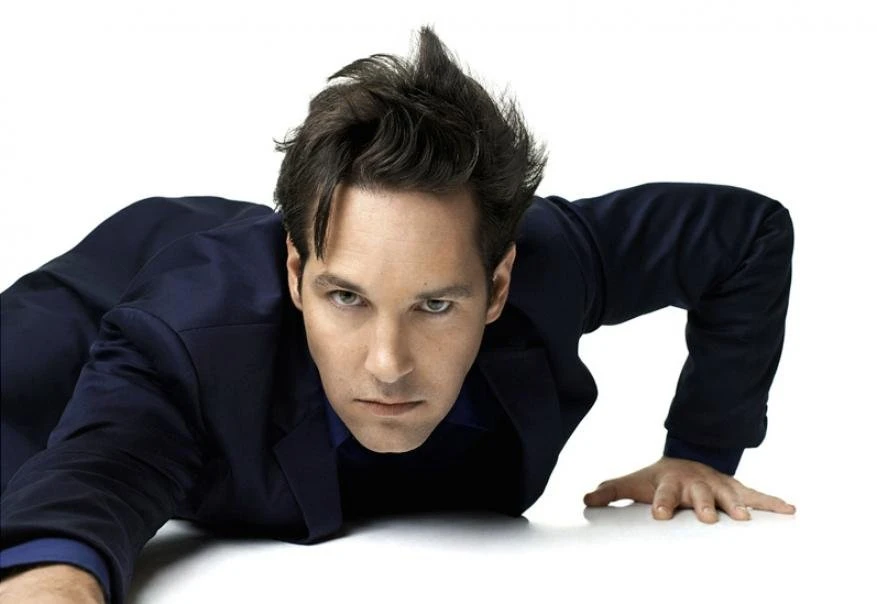 Actor Paul Rudd lies prone on a white surface in a navy suit jacket with an intense gaze directed at the camera in a portrait shot.