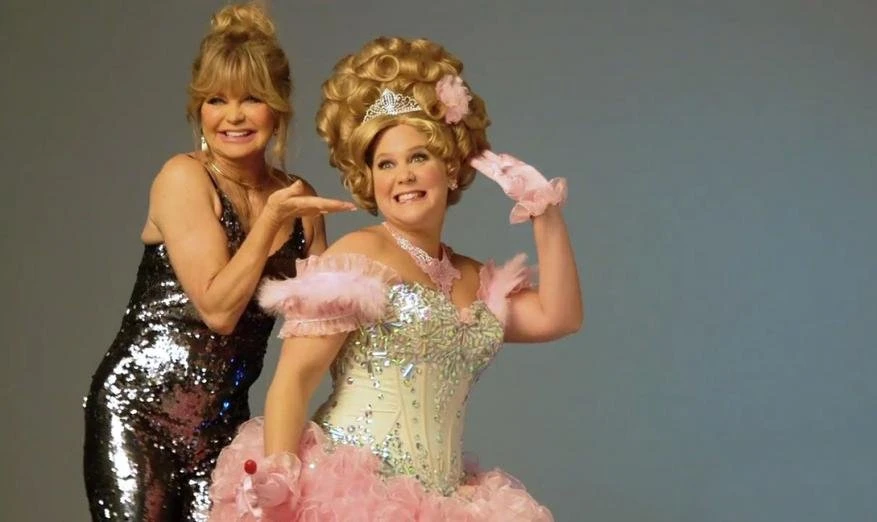 Two women pose in a photo studio, one in a sparkly dress, the other in a princess costume, against a plain grey backdrop.