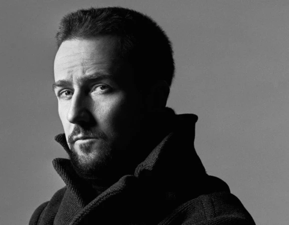 Monochrome portrait of a man with short hair wearing a high-collared dark coat against a grey studio backdrop.