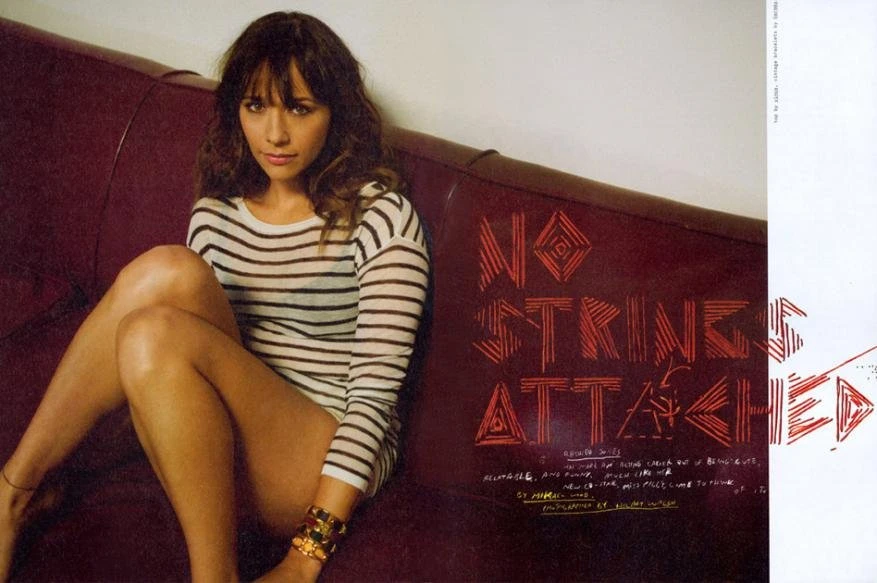 Rashida Jones is seen indoors, wearing a striped shirt and bracelets. She is seated in a reclined position in front of a wall with graphic text.