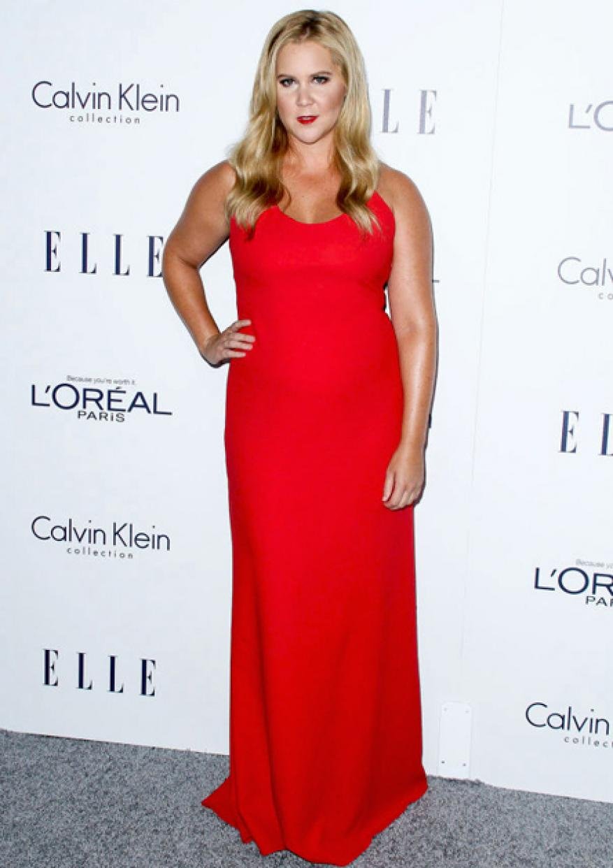Amy Schumer poses on a grey carpet in a long red dress at a Calvin Klein and Elle event, smiling at the camera with blond hair.