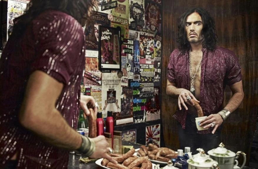 Adult male stands in front of a mirror, holding a sausage and a cup; kitchen with food, plates and bottles; wall covered in posters.