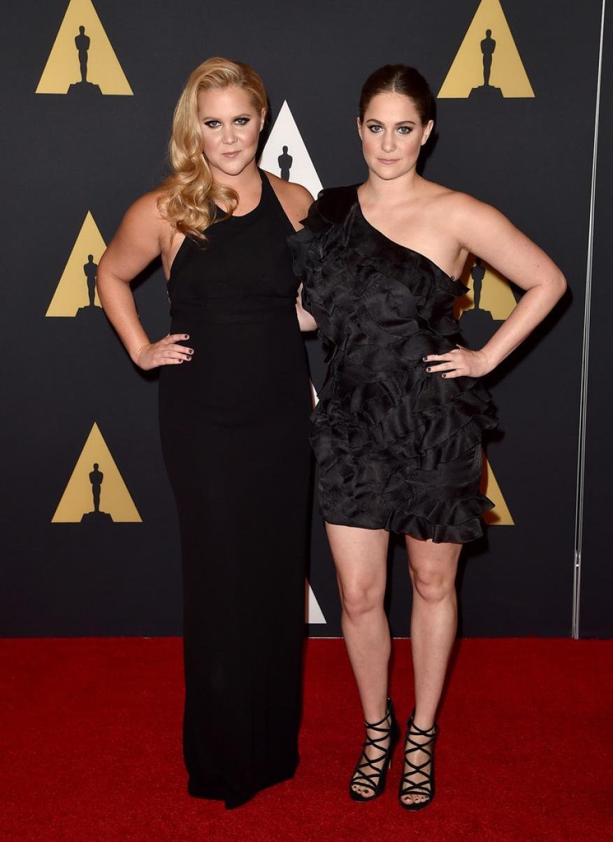Two adult women pose on a red carpet in black formal dresses. They are at an award ceremony with backdrop logos.