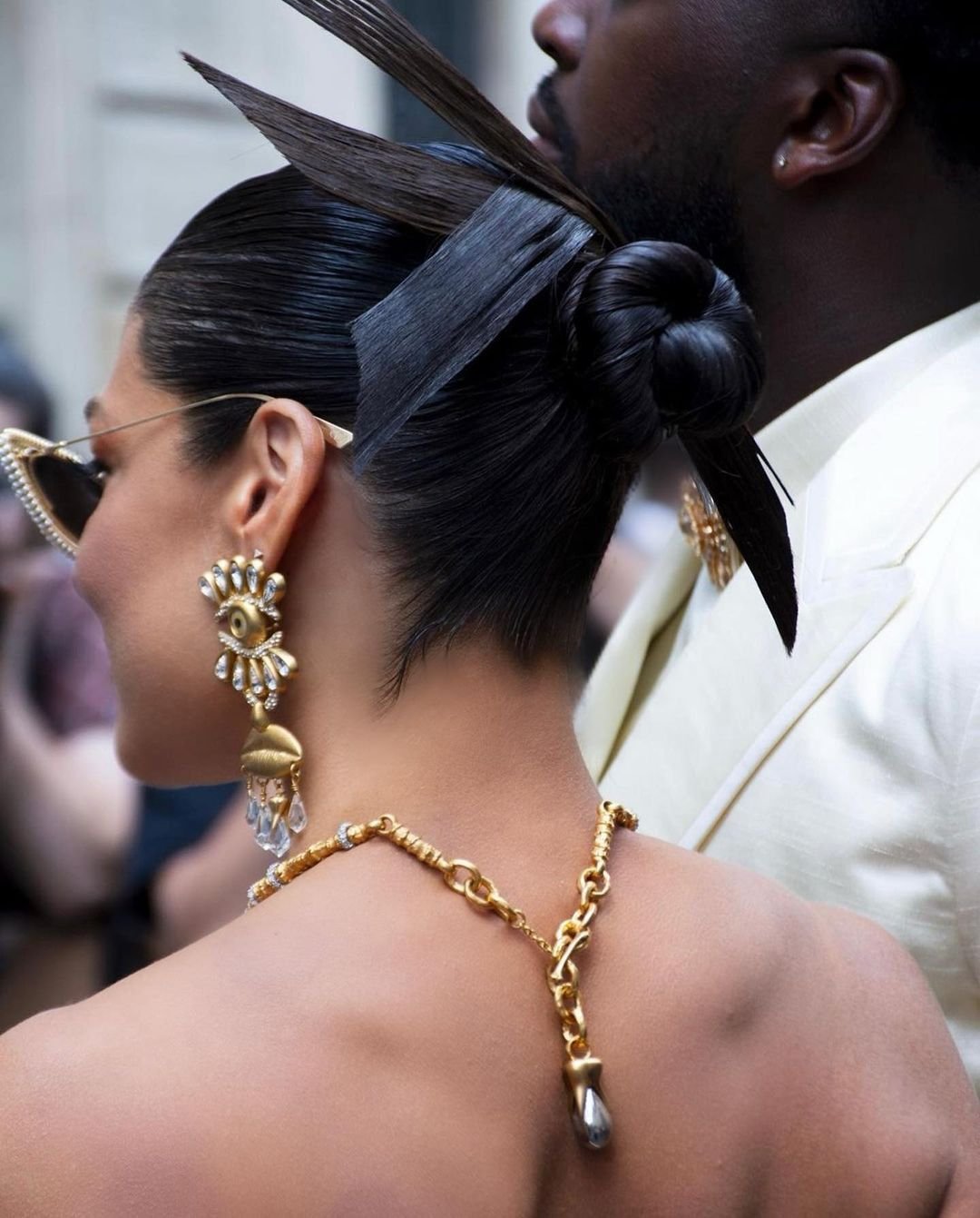 Close-up of a woman with a creative updo, statement earrings, and a gold necklace, standing near a man in a white suit. Fashion.