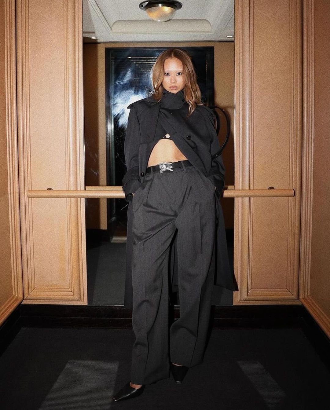 Adult female fashion model in black coat and trousers posing indoors with neutral wall and doorway background under overhead lighting.