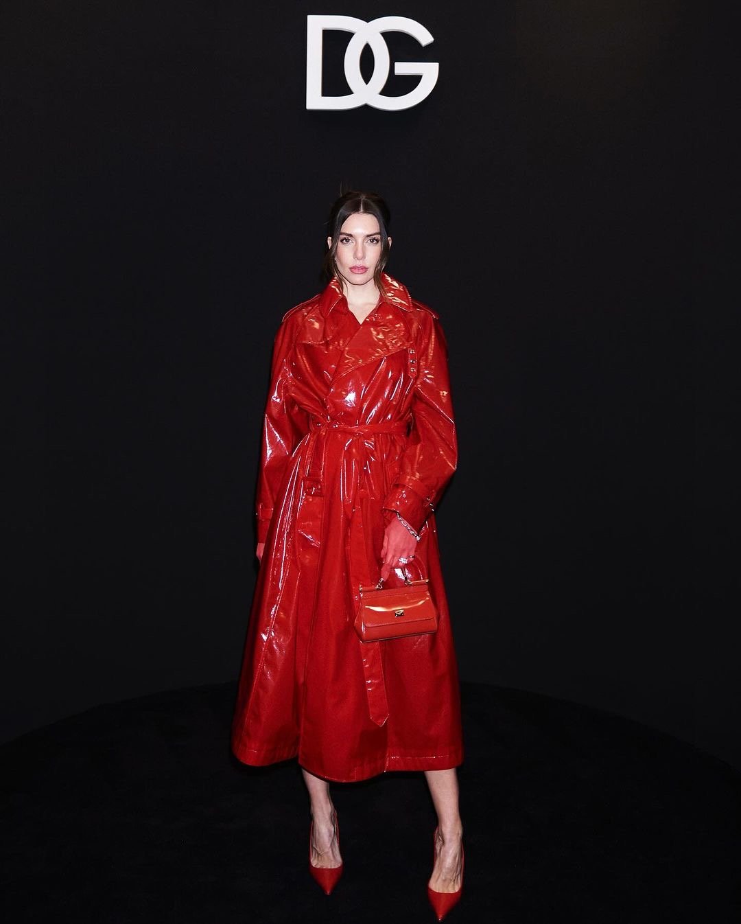Adult female wearing red patent leather trench coat with matching purse and high heels posing in front of a black background and DG logo.