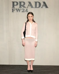 A woman wearing a pink knit cardigan and matching skirt stands against a grey wall with PRADA FW24 branding.