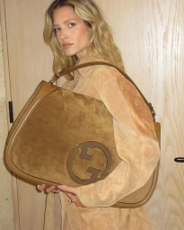 Woman holding a large brown suede shoulder bag featuring a prominent Gucci logo in a room with light wood paneling.
