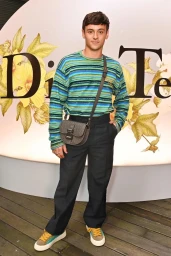 Tom Daley wearing a striped blue sweater, dark trousers, and a crossbody bag standing in front of a floral backdrop.