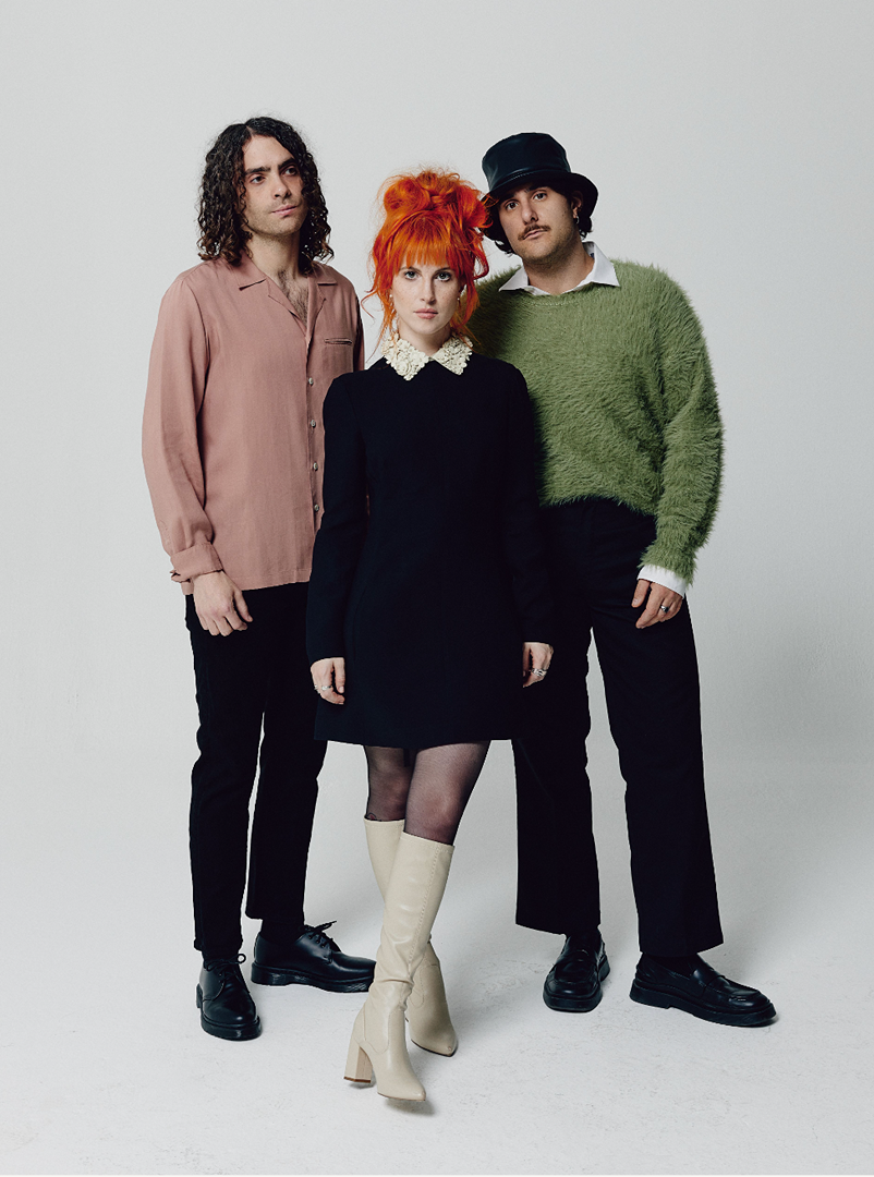 Three band members stand in a photo studio. The woman in the center has orange hair, wearing a black dress and cream boots.