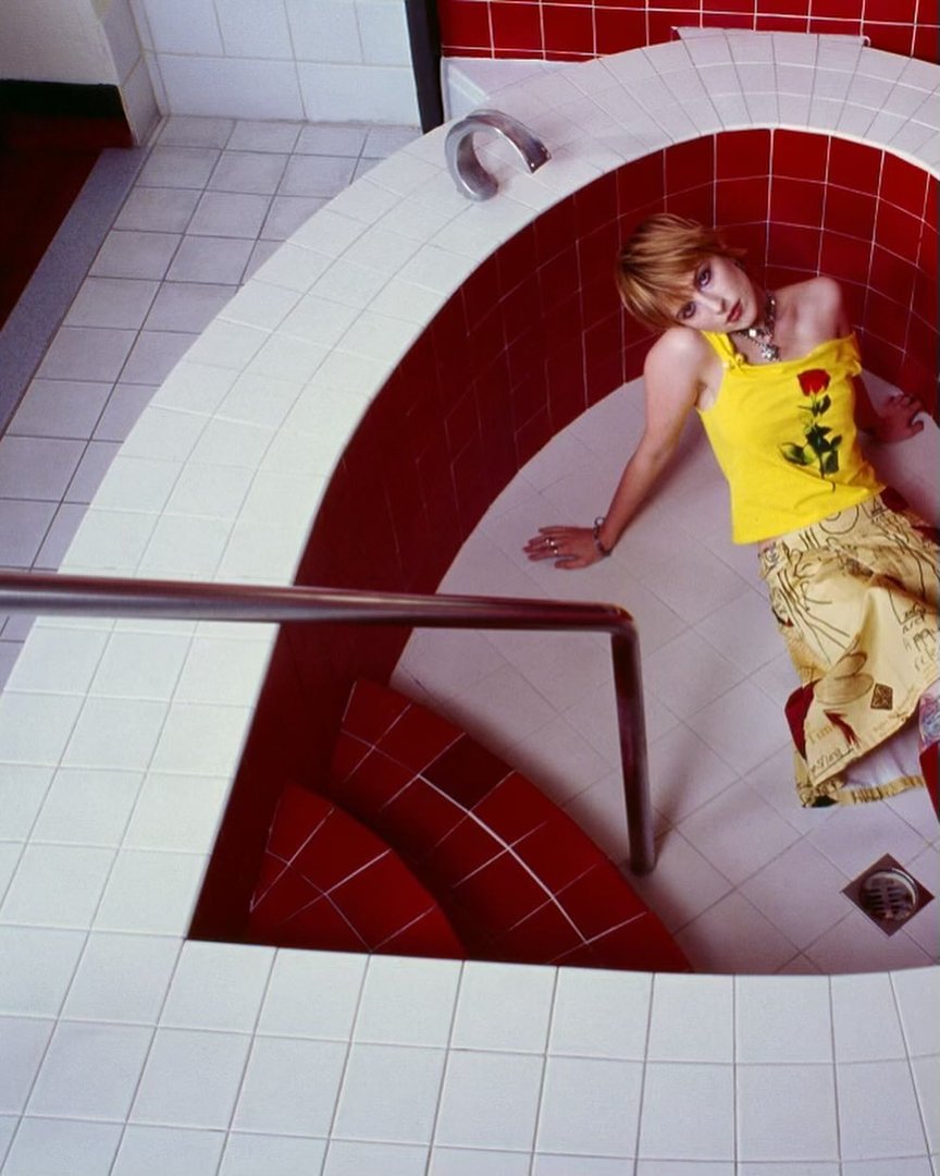 Indoor portrait shot of a woman with blonde hair reclining in a sunken tub, wearing a yellow top and patterned skirt in a tiled room.