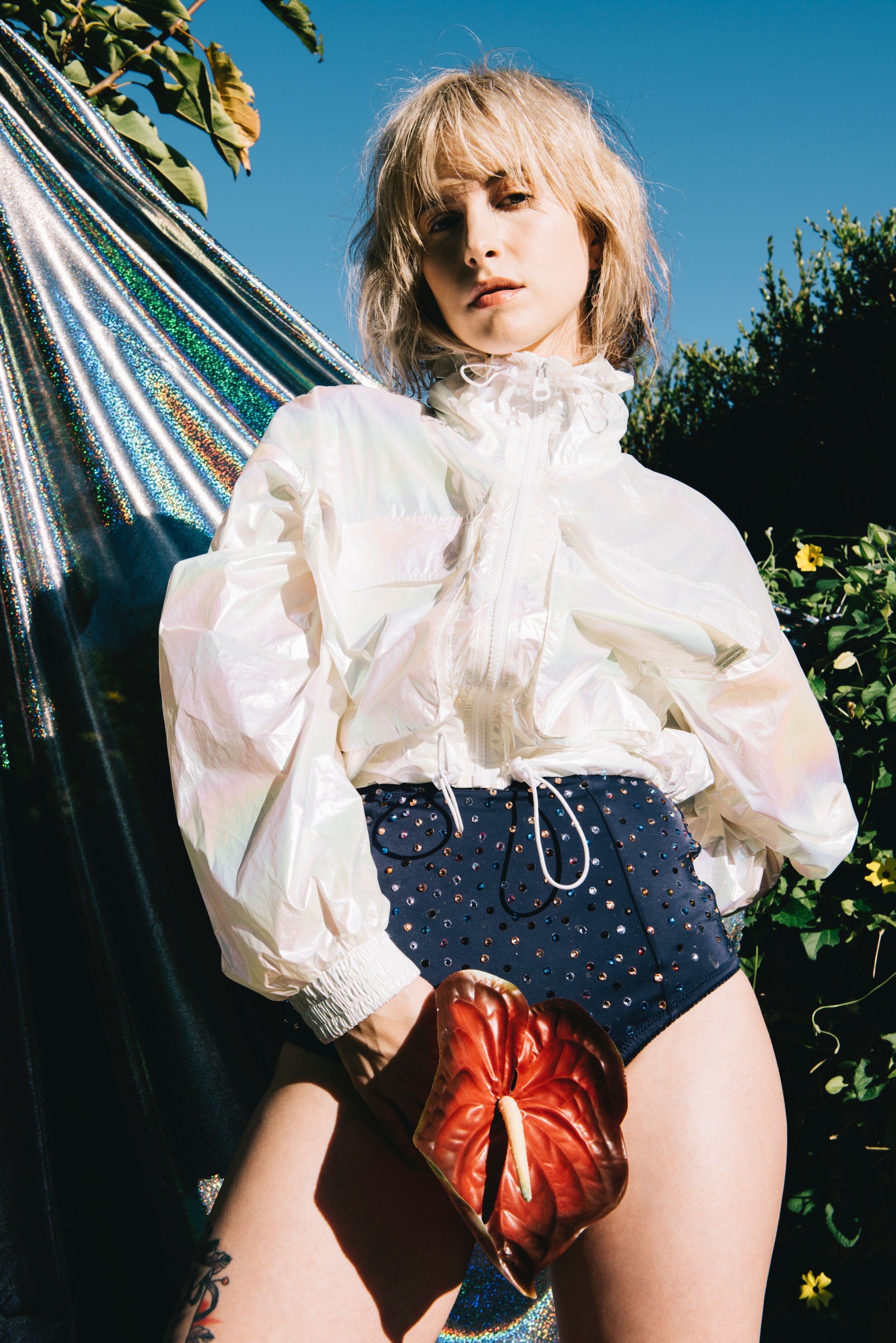 Adult female poses outdoors wearing a white jacket, dark shorts, and holds a red flower against a metallic and foliage background.