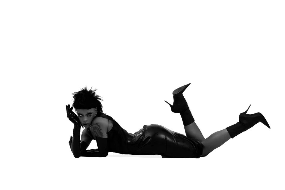 Adult female model lying down in a black dress and heels against a stark white background in a studio fashion photography shot.