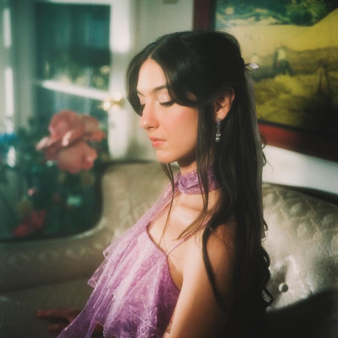 Brunette woman in a lavender dress sits on a couch, gazing downward with a soft, filtered light highlighting her elegant pose.