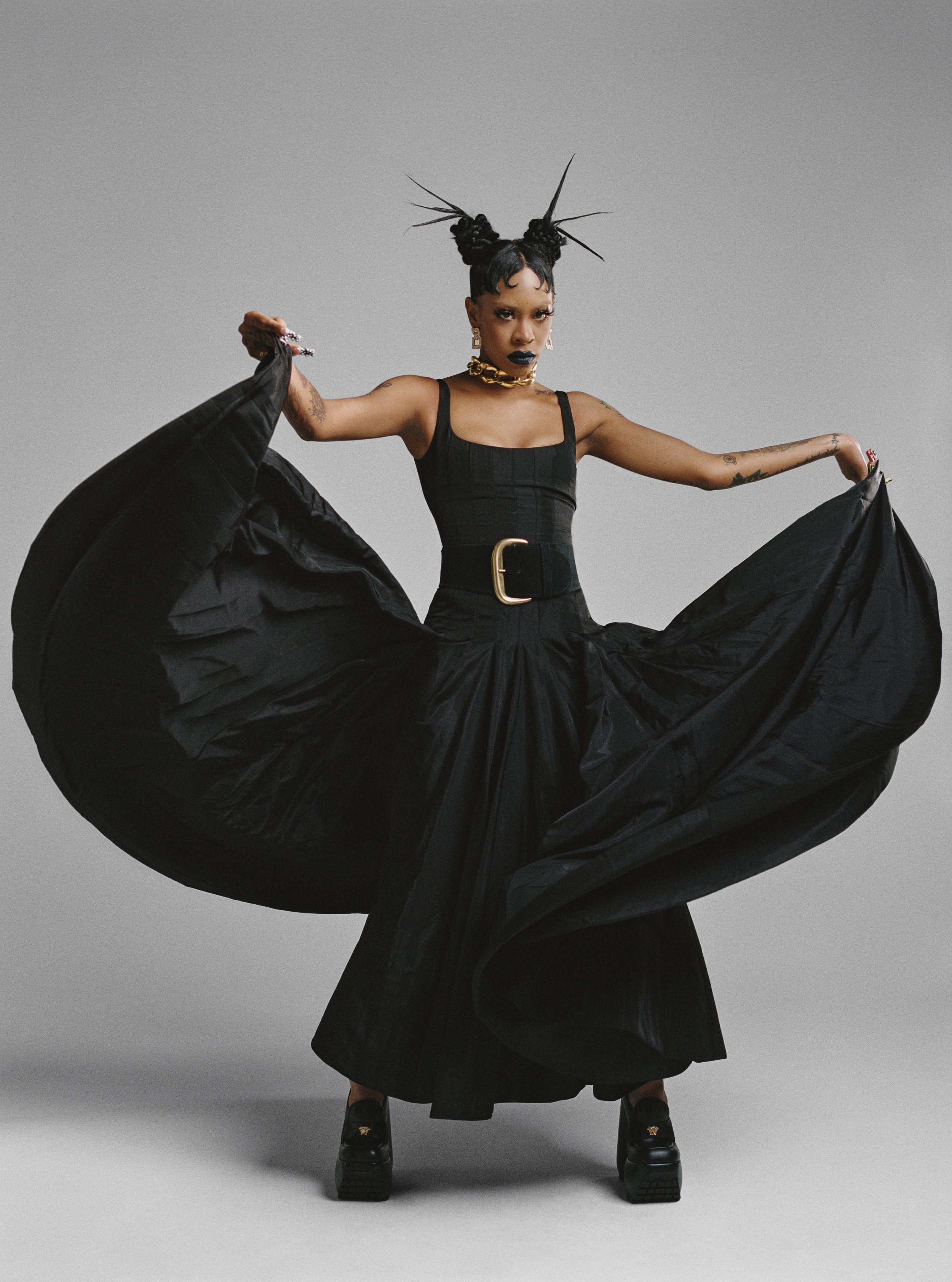 A person posing in a black dress with a full skirt, accessorized with a gold chain and belt, in a fashion studio setting.
