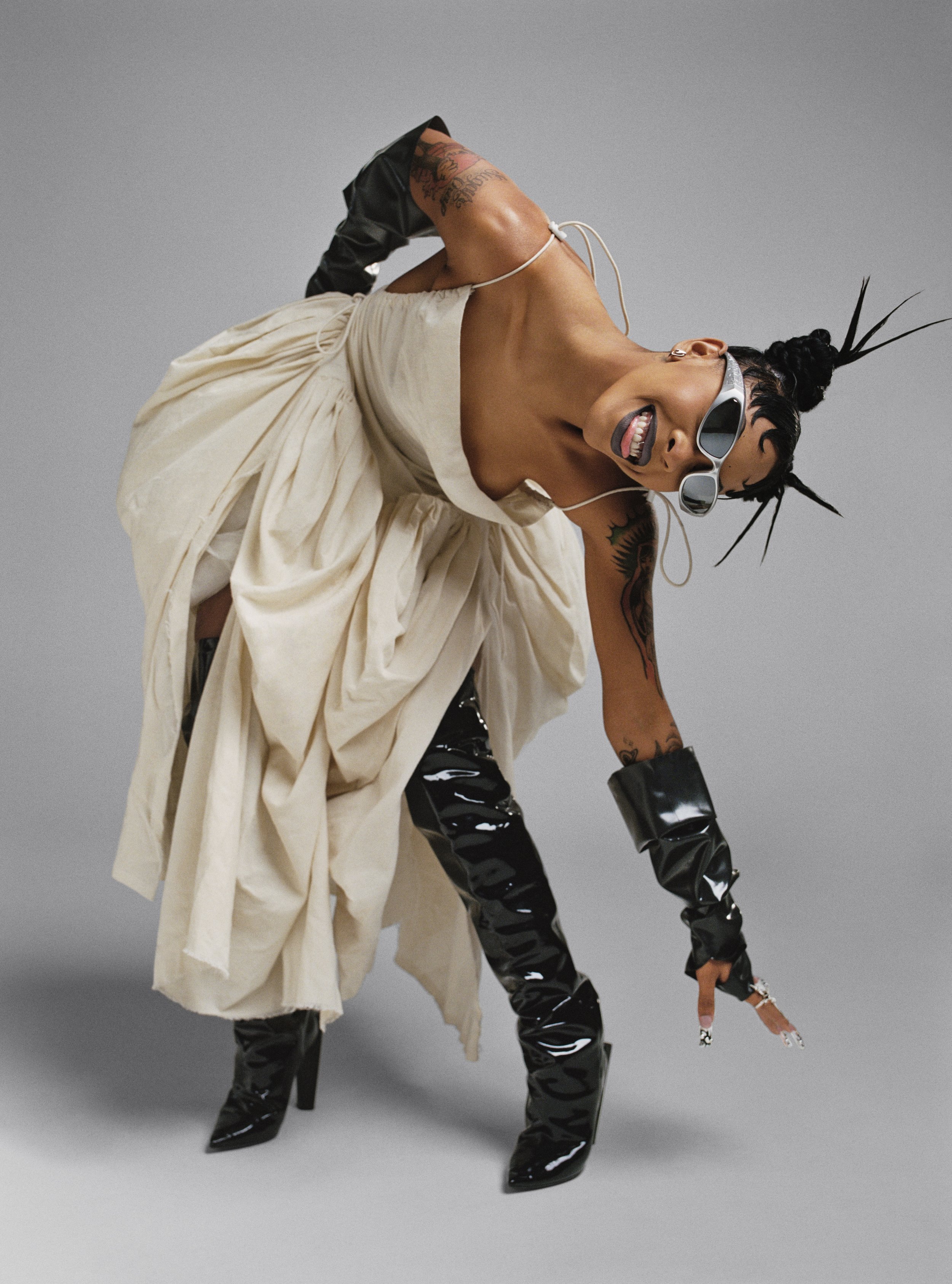 A woman in a light-colored dress, black boots, and sunglasses poses against a grey background in a fashion or editorial shot.