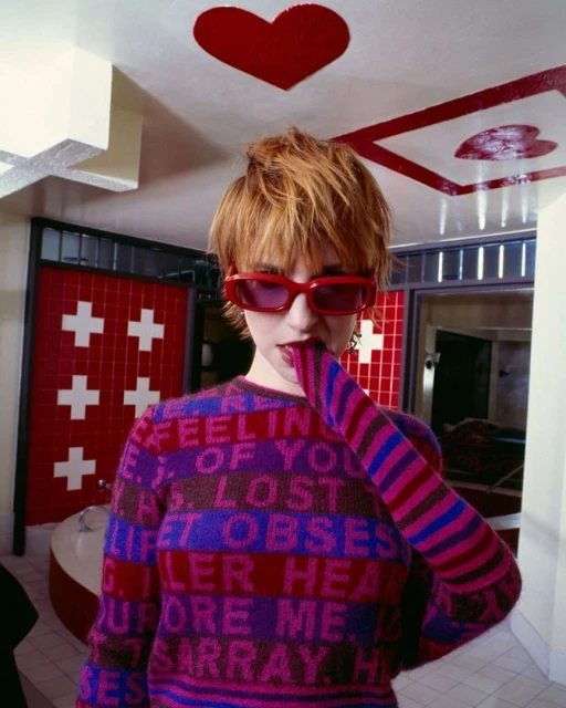 Portrait of a person with short hair and red glasses wearing a sweater with writing in an indoor setting with a white and red background.