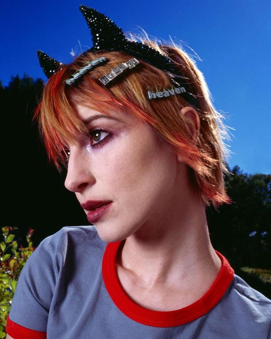 Woman with orange hair and 'heaven' hair clip wearing black studded devil horn headband against a blue sky with blurred green trees.