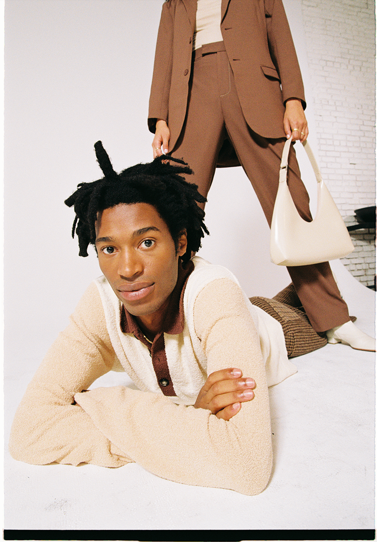 Fashionable studio shot of two African-descent adults wearing complementary beige and brown outfits with a handbag accessory.