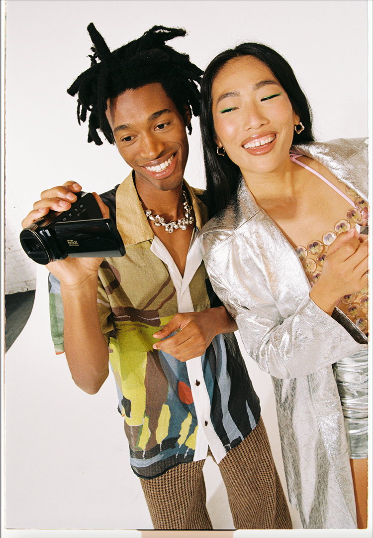 A stylish couple poses in front of a white background, one holding a camcorder, both smiling, wearing fashionable clothing and jewelry.