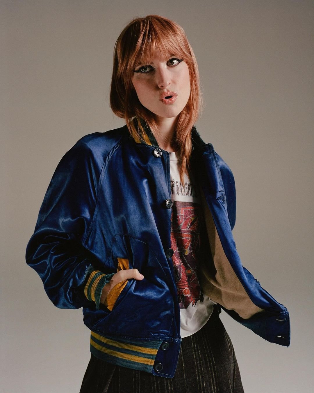 Portrait of a young woman with red hair wearing a blue bomber jacket and t-shirt in a studio setting, looking at the camera.