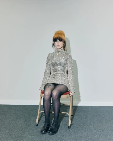 A woman with orange hair wearing a glittering dress, black tights, and shoes sits on a chair in front of a white wall and grey floor.