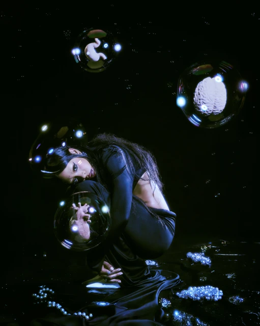 Woman in dark clothing squats surrounded by illuminated spheres in a dark space, creating a surreal and artistic scene with mysterious lighting.