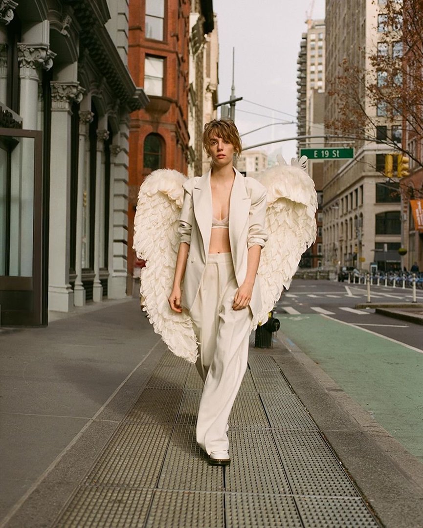 Adult female in a white suit and angel wings walking down a sidewalk in a city with buildings and a street sign visible.