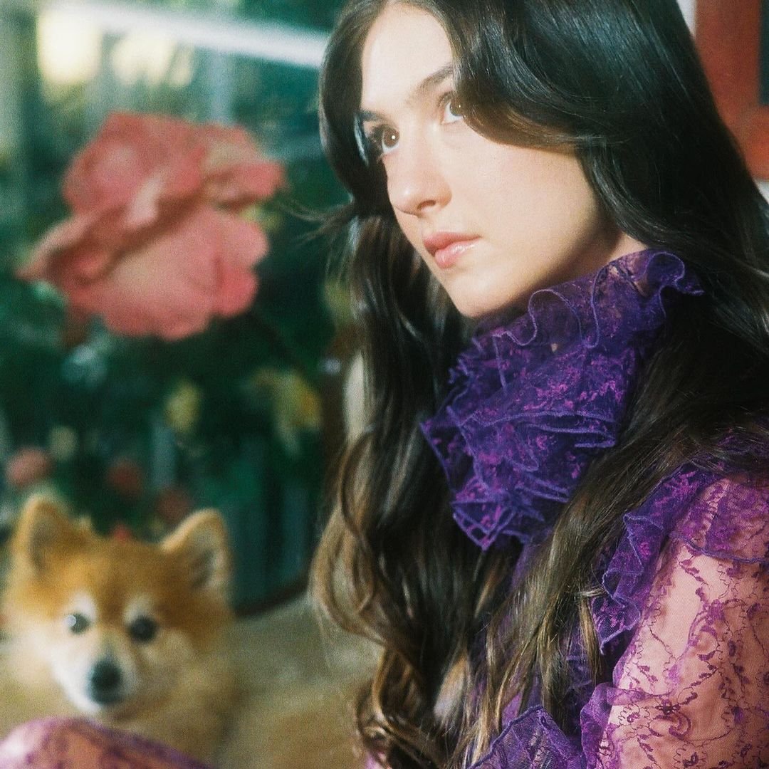 Portrait of a woman wearing a purple lace ruffled blouse with a small dog and flowers in the blurred background.