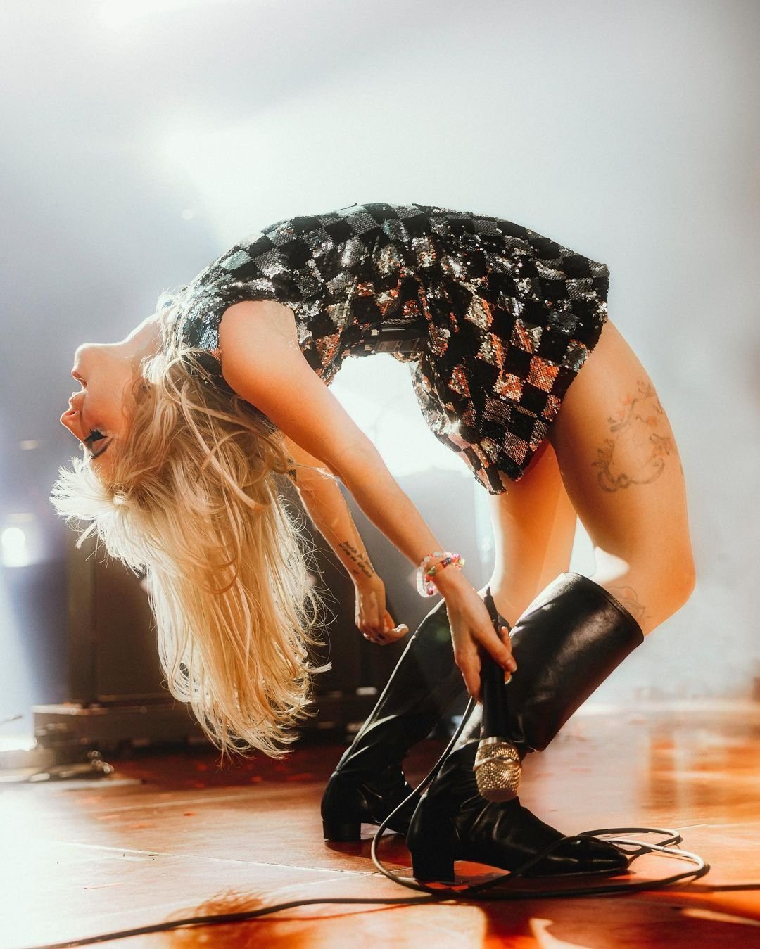 Dynamic full shot of a singer with long blonde hair, wearing a sequined dress and high black boots, bending backwards on stage.