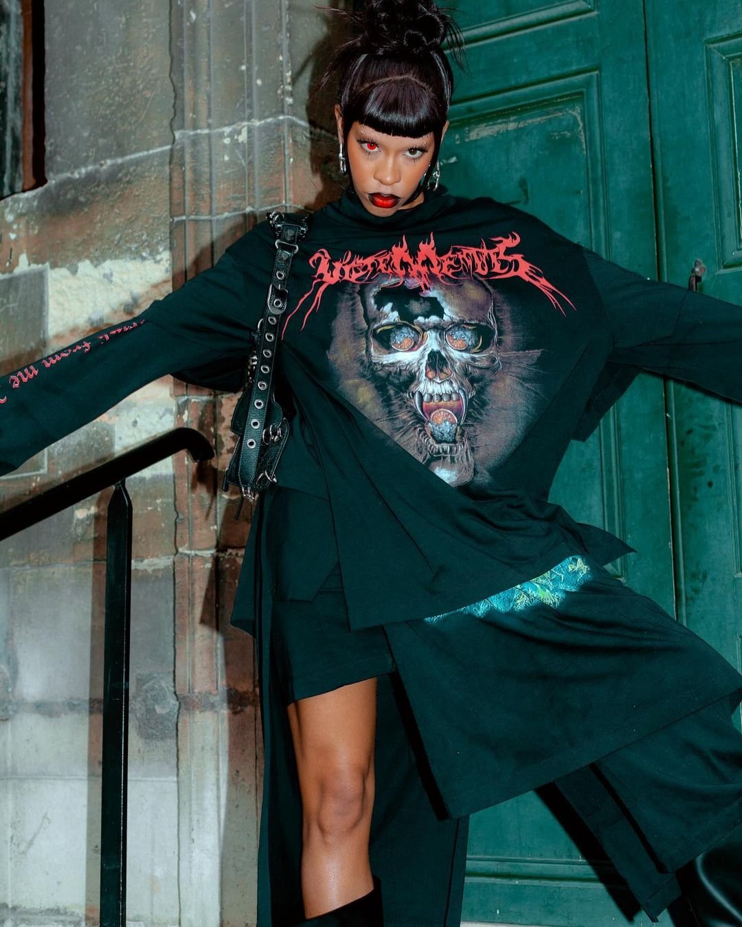 Woman in Vetements oversized skull graphic tee and black skirt poses in front of a stone building, accented by a black studded purse.