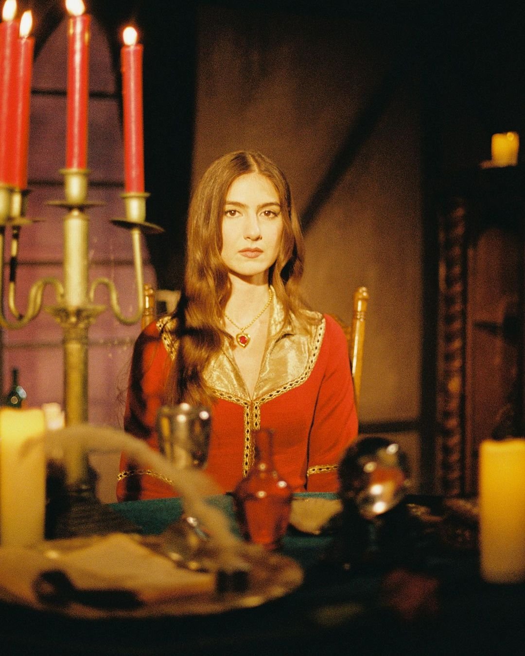 Portrait of a woman with long brown hair wearing a red dress with gold trim sitting at a table with candlesticks and objects.