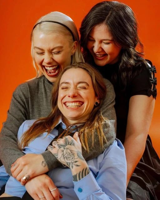 Three adult women are tightly embraced, laughing joyfully with an orange backdrop. They are dressed casually in muted tones.