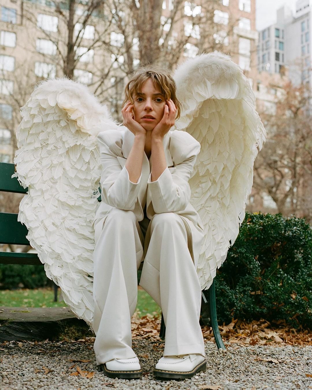 Adult female model with angel wings and a white suit sits on a park bench surrounded by trees and buildings in an outdoor setting.