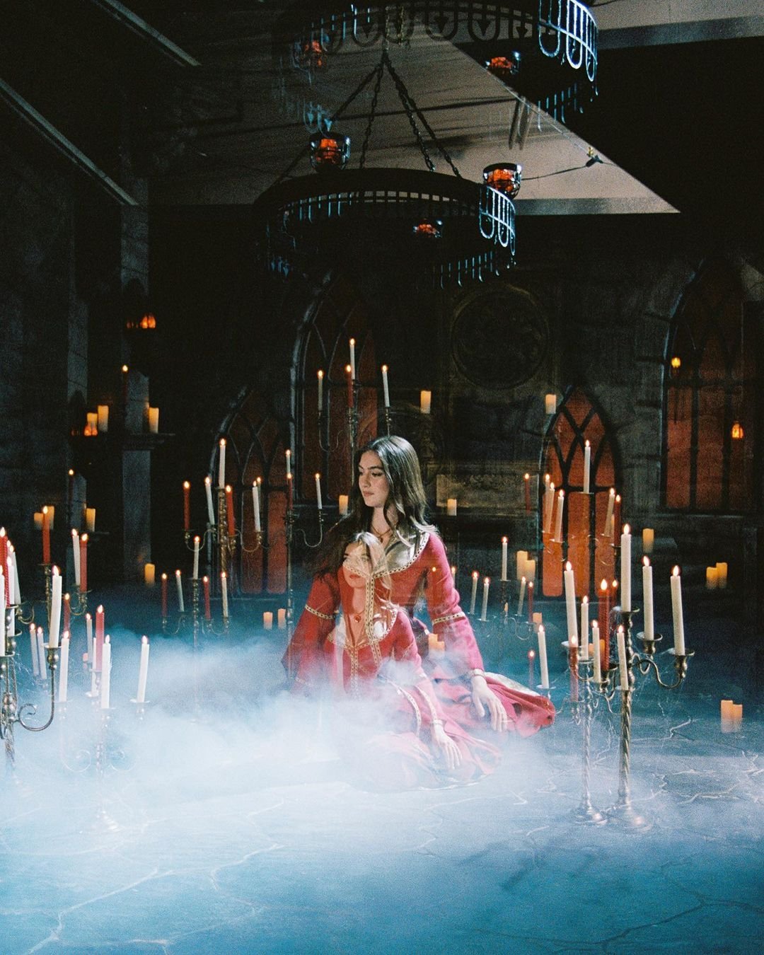 Two women in red gowns amidst a sea of candles, set in a dark stone room, evoke a scene of gothic elegance and mystery.