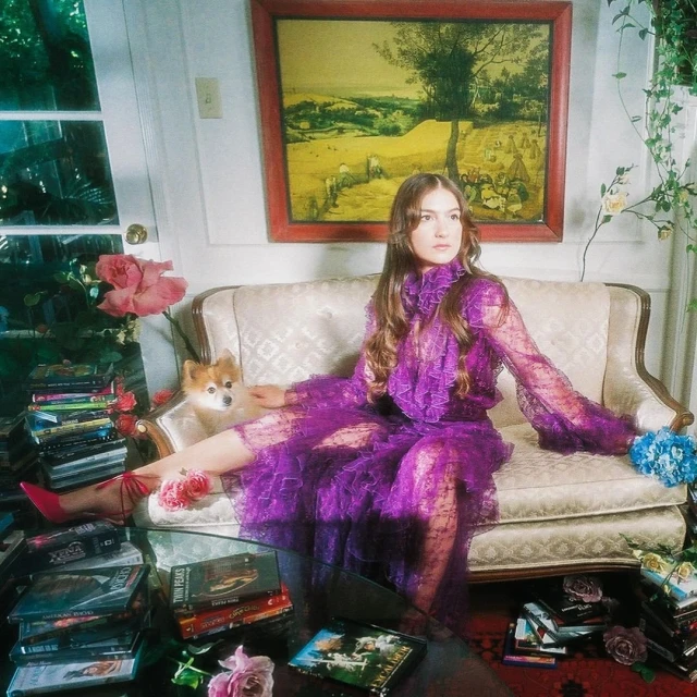 Adult female in an elaborate purple dress sitting on a vintage couch with a dog, surrounded by roses and a collection of DVD cases.