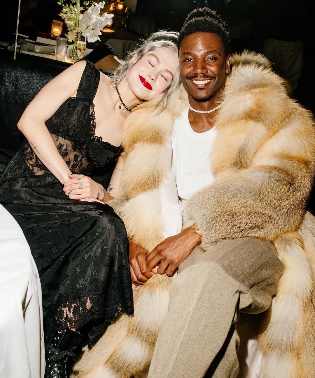 Smiling man in fur coat with woman in black lace dress. Indoor image featuring two adults, neutral tones, and soft lighting.