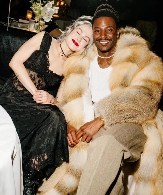 Smiling man in fur coat with woman in black lace dress. Indoor image featuring two adults, neutral tones, and soft lighting.