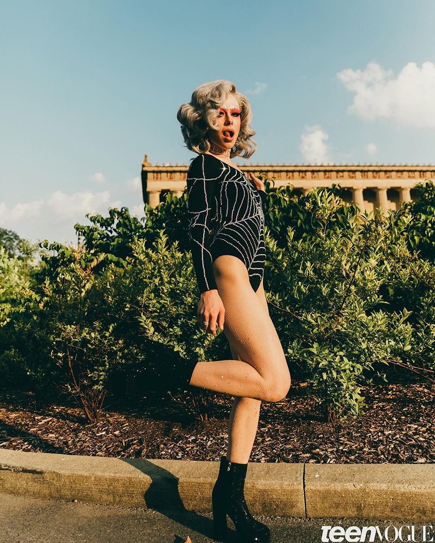Person in a patterned bodysuit and platform boots poses outdoors in front of green shrubs and an old building on a sunny day.