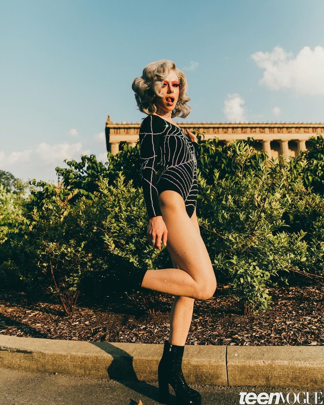 A person in a black bodysuit and glitter boots poses outdoors with a building in the background in a bright sunny day.