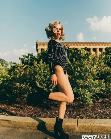 A person in a black bodysuit and glitter boots poses outdoors with a building in the background in a bright sunny day.
