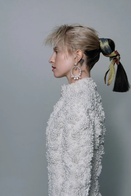Profile view of a woman with blonde hair in a high ponytail, wearing a pearl-beaded dress and pearl earrings against a gray backdrop.