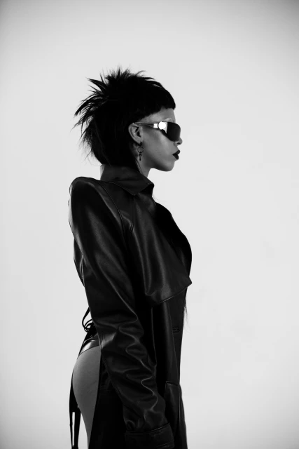 Profile shot of a woman with a mohawk and sunglasses wearing a leather trench coat against a white backdrop in a studio setting.