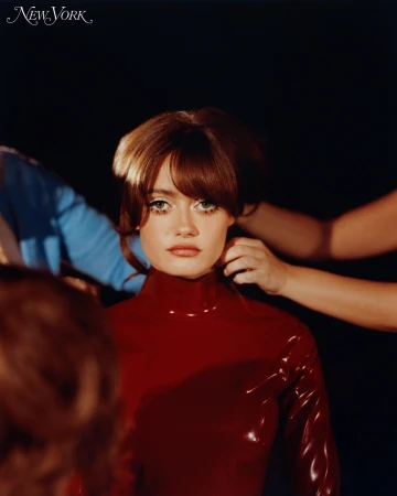 Portrait of a woman with auburn hair wearing a red turtleneck, positioned against a dark background, with hands adjusting her hair.