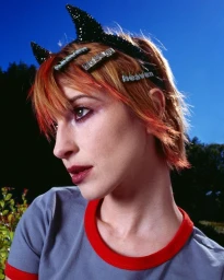 Close-up profile of a woman with red hair wearing sparkling devil horns headband and text hair clips against a clear blue sky