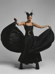 Black woman with elaborate hair styling holding out the wide skirt of her black gown against a seamless grey studio backdrop