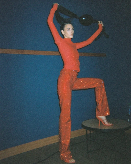 A woman in a red outfit poses on a table against a dark blue wall, showcasing a fashion-forward look with high heels.