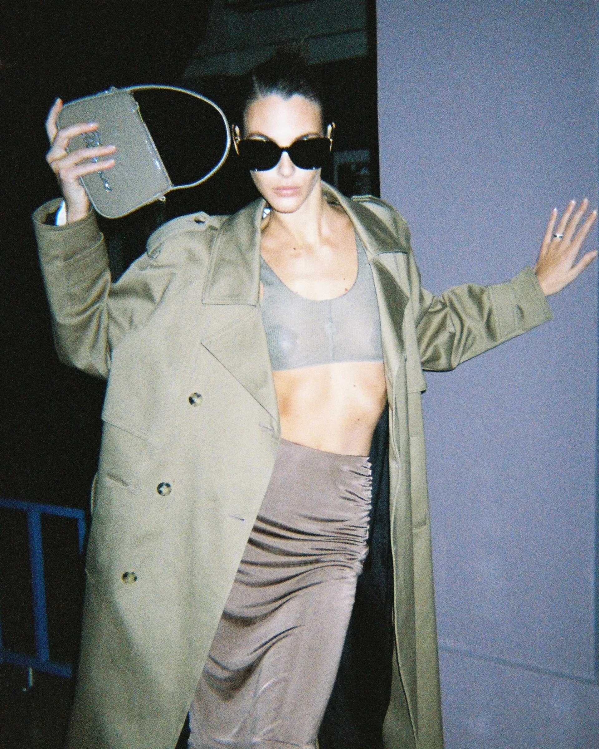Adult female wearing a trench coat and sunglasses poses with a purse, showcasing fashion apparel and accessorizing with jewelry.