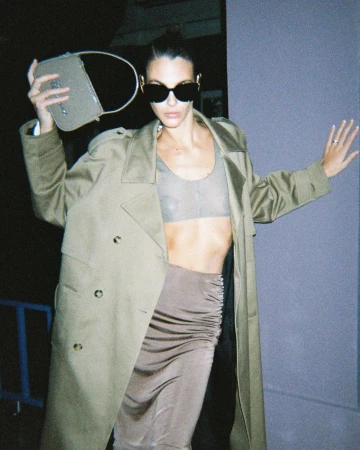 Adult female wearing a trench coat and sunglasses poses with a purse, showcasing fashion apparel and accessorizing with jewelry.